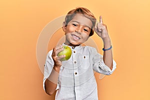 Adorable latin kid holding green apple smiling with an idea or question pointing finger with happy face, number one