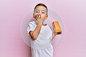 Adorable latin toddler holding gold ingot covering mouth with hand, shocked and afraid for mistake