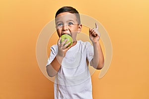 Adorable latin toddler smiling happy eating green apple looking to the camera over isolated yellow background photo