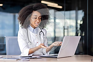 Adorable latino woman in white lab coat and glasses sitting at desk in medical center and having online conference on