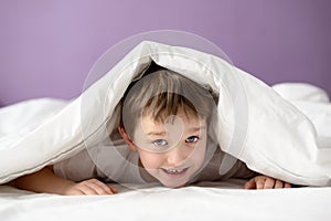 Adorable laughing boy playing in bed under a white blanket or coverlet
