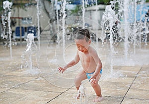 Adorable little Asian baby boy having fun on water stream of a sprinkler. Kid playing in playground fountain in aqua park