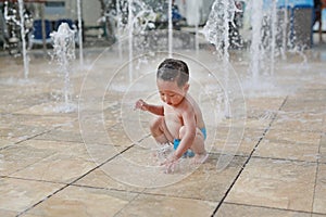 Adorable little Asian baby boy having fun on water stream of a sprinkler. Kid playing in playground fountain in aqua park