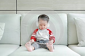 Adorable little Asian baby boy in headphones is using a smartphone lying on the sofa at home. Child listening to music on