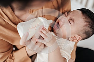 Adorable little Asian baby uncomfortable crying during drinking from baby bottle. Mother holding adorable her son with milk