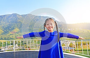 Adorable little Asian child girl feeling free with arms wide open at beautiful trees and mountains on blue sky