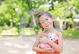 Adorable little Asian child girl hugging teddy bear doll in the garden with looking at camera. Close up happy kid in summer park