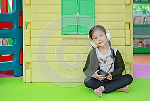 Adorable little Asian child girl listening music by headphone near toy playhouse in playground