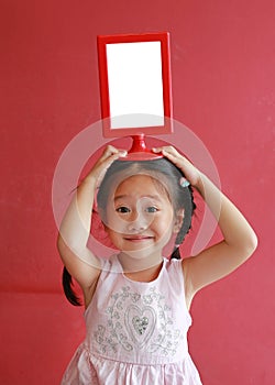 Adorable little asian girl holding empty whiteboard on her head against red wall background