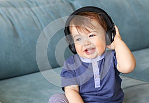 Adorable little baby boy listening to music in headphones on his head sitting on blue sofa at home