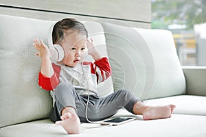 Adorable little baby boy sitting on sofa and listening music at headphones by smartphone. Kid using earphones to relaxing indoors