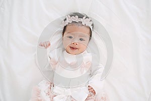 Adorable little baby girl in a beautiful dress with flower band lying on a white blanket.