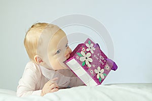 Adorable little baby lying on the white blanket and holding purple gift bag in his hands. Horisontal studio shot. Happy