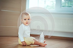 Adorable little baby sitting in bedroom on the floor with bottle with milk or water and laughing. Infant Childhood Kids People