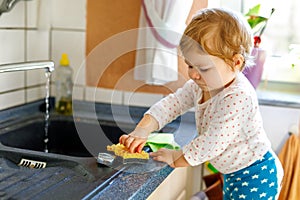 Adorable little blond baby girl washing dishes in domestic kitchen.