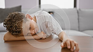 Adorable little blond boy, with a sad expression, comfortably leaning on a living room table