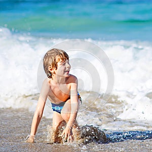 Adorable little blond kid boy having fun on ocean beach. Excited child playing with waves, swimming, splashing and happy