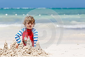 Adorable little blond kid boy having fun on tropical beach of carribean island. Excited child playing and building sand