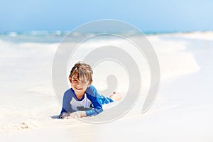 Adorable little blond kid boy having fun on tropical beach of carribean island. Excited child playing and surfing in sun