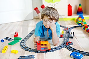 Adorable little blond kid boy playing with colorful plastic blocks and creating train station. Child having fun with