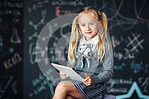 Adorable little blond schoolgirl in school uniform holding white digital tablet. Chalkboard with school formulas background. Back