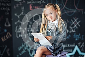 Adorable little blond schoolgirl in school uniform holding white digital tablet. Chalkboard with school formulas background. Back