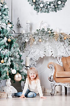 Adorable little blonde girl in a blue jeans sitting on a floor near christmas tree and looking on toy.