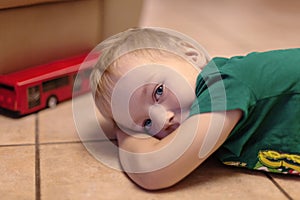 Adorable little boy with blue eyes lays on the ceramic floor with toy red bus. Blonde hair, green t-shirt.