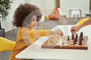 Adorable little boy making a move, playing chess with adult while sitting at the table indoors