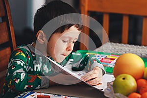 Adorable little boy opens page of exercise book. Cute boy reading book on chair at home. Happy child reading a book. School &