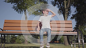 Adorable little boy sitting on the bench in the park. Cute child spending time alone outdoors. Summertime leisure