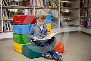 Adorable little boy, sitting in library, reading book and choosing what to lend, kid in book store