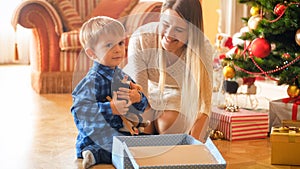 Adorable little boy sitting under Christmas tree and hugging plush toy he received in gift box from Santa