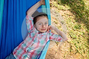 Adorable little child girl smiles at camera while relaxing on blue hammock in the backyard, enjoying a weekend outdoors