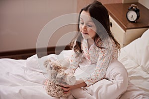Adorable little child girl in stylish pajamas, sitting on bed and playing with her plush toy sheep before going to bed