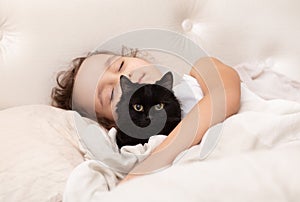 Adorable little cute girl child is sleeping in the bed with her pet black cat. The girl is hugging the teddy bear