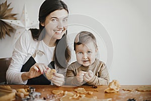Adorable little daughter with mother making together christmas gingerbread cookies on messy wooden table. Cute toddler girl helps