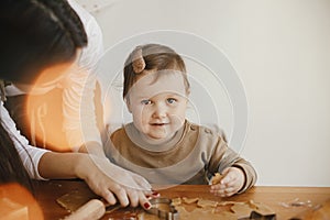 Adorable little daughter and mother making together gingerbread cookies on messy wooden table in modern room. Cute toddler girl