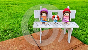 Adorable little dog and beautiful girl dolls sitting on the white chair on the green grass garden look lovely and cute.