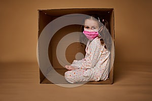 Adorable little European girl with two ponytails in pajamas wearing a pink medical protective mask sits inside a cardboard box,