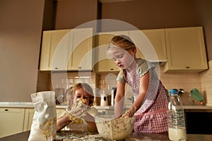 Adorable little girl in apron kneading dough while making cookies together with her brother on the kitchen table at home