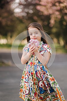 Adorable little girl in beautiful dress hold flowers in hand. Sakura tree in the park. Female kid model