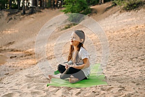 Adorable little girl with brown long hair sitting on green mat on sandy beach with bottle of water in hand and smiling.