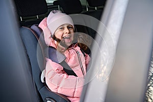 Adorable little girl, cheerful child in pink warm clothes smiling sitting fastened with a secure seat belt in the modern car