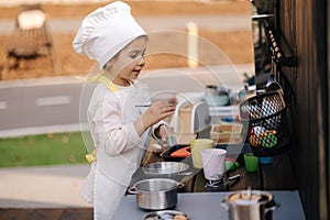 Adorable little girl in chef's coat and cap cooks at the children's toy kitchen. Playing on little kids city