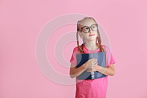 Adorable little girl in glasses with funny pigtails is holding book, smiling looking up is thinking on pink background