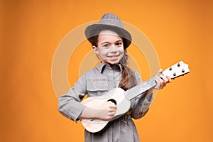 Adorable little girl in a gray hat smiles and plays the guitar while standing on an orange background