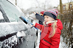 Adorable little girl helping to brush a snow from a car. Mommy`s little helper.