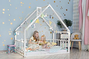Adorable little girl and her young mother are reading a book and smiling while sitting in decorated house bed