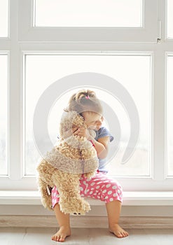 Adorable little girl hugging a teddy bear. Cute baby at home in white room is sitting near window.